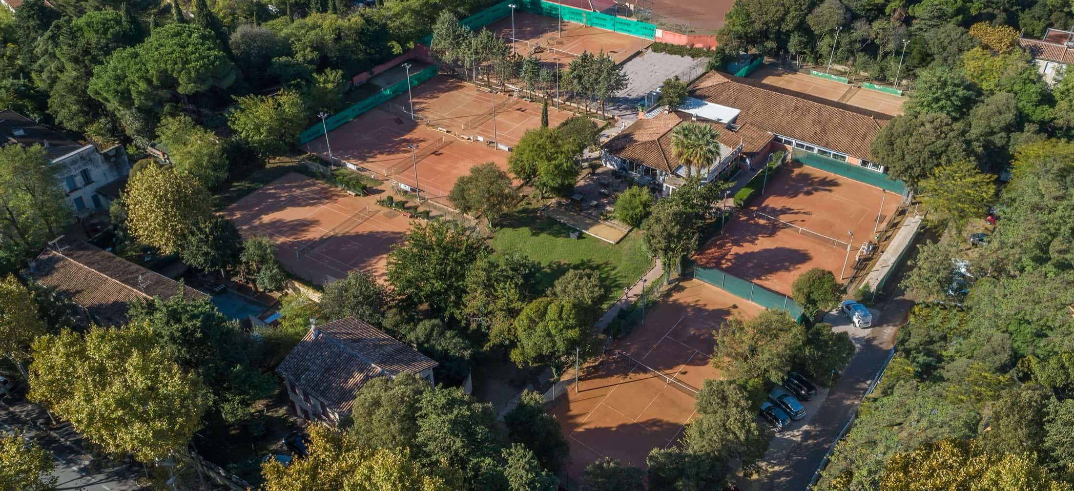 Vue aérienne des courts en terre battue du Tennis Club de la Pierre Rouge, entourés de pins, avec le club-house au centre